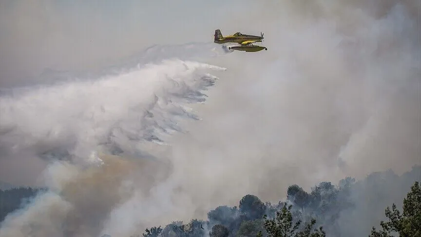 A yellow single-engine firefighting plane banks over a smoking hillside, trailing a long white curtain of water above burning pine forest.